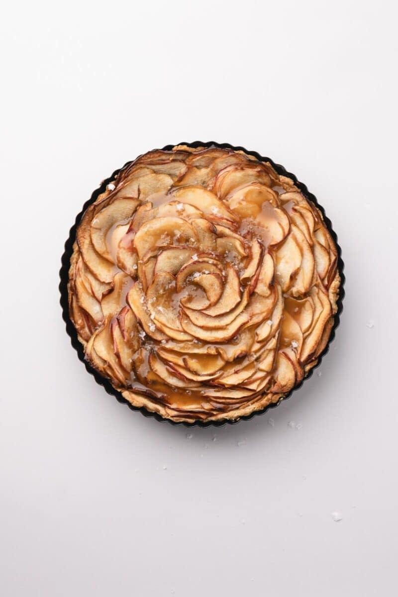 A round caramel apple tart with thinly sliced apples arranged in a spiral pattern and drizzled with caramel glaze, in a black tart pan on a white background.