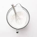 Clear glass mixing bowl with flour and a metal whisk inside, viewed from above on a white surface.