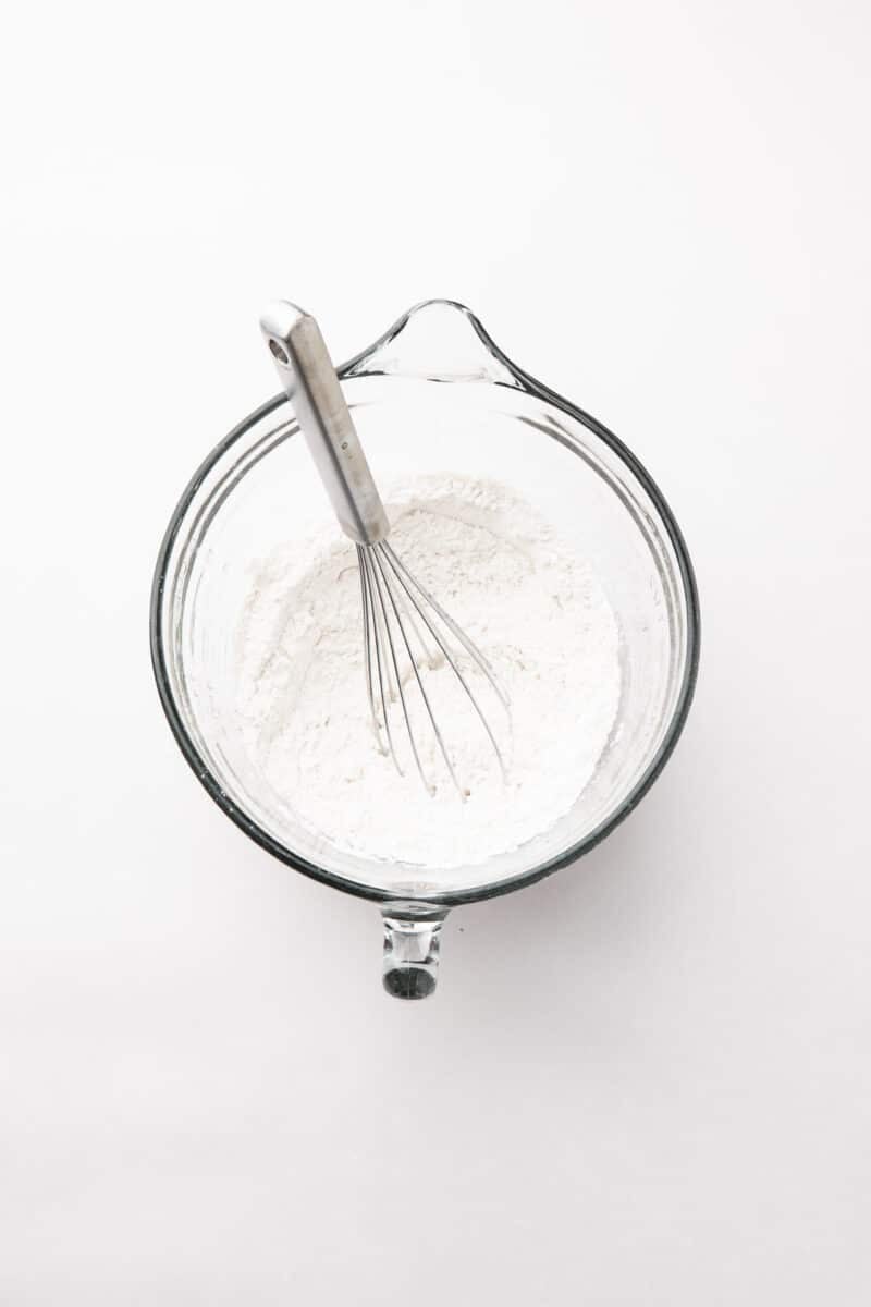 Clear glass mixing bowl with flour and a metal whisk inside, viewed from above on a white surface.