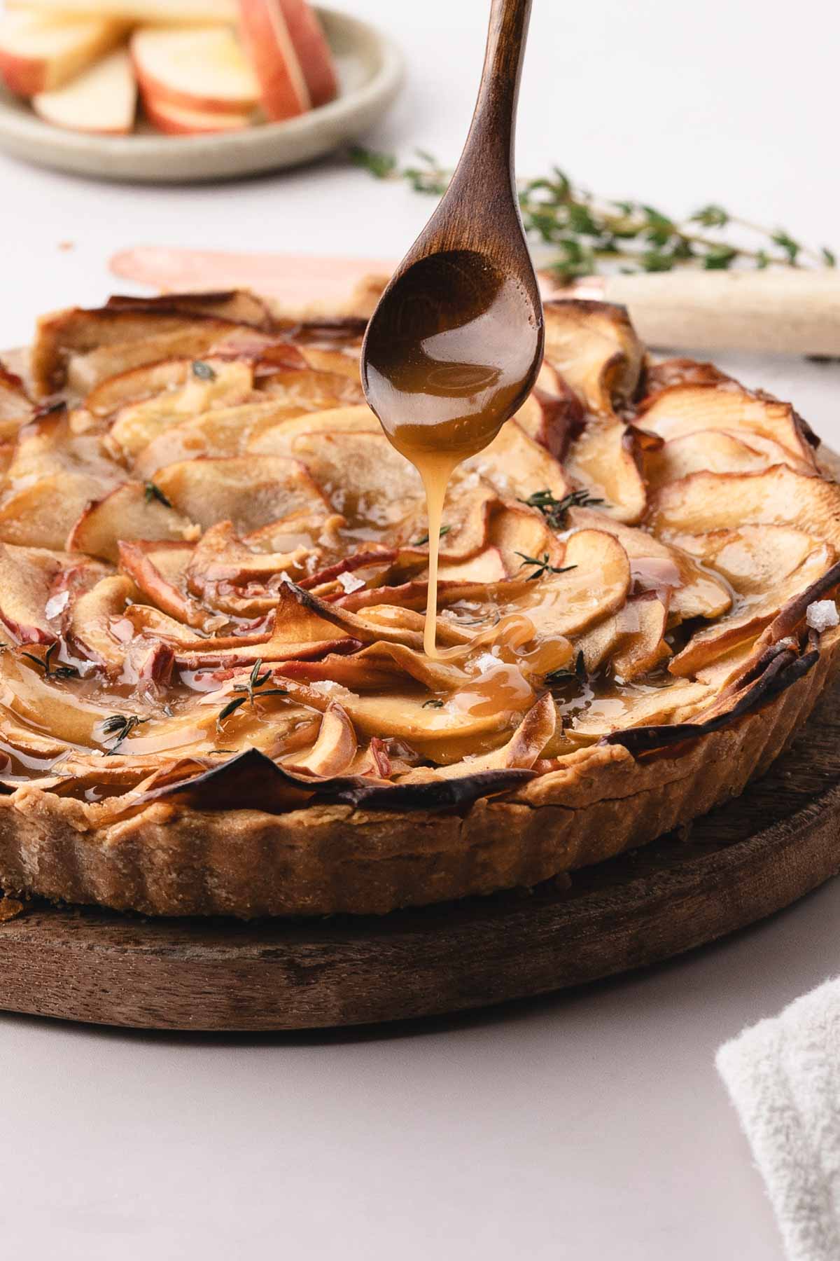 A wooden spoon drizzles caramel sauce over a baked apple tart on a wooden board, with apple slices and herbs visible in the background.