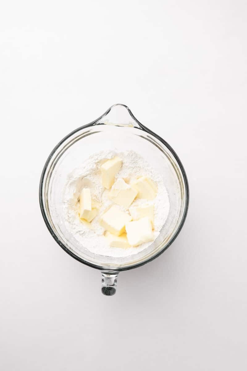 A glass mixing bowl with flour and several pieces of cubed butter on a white surface.