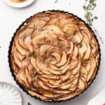 A caramel apple tart with thinly sliced apples arranged in a spiral, drizzled with caramel sauce, garnished with herbs, and surrounded by cinnamon sticks, apple slices, and a towel.