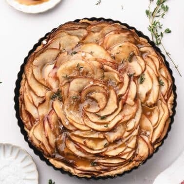 A caramel apple tart with thinly sliced apples arranged in a spiral, drizzled with caramel sauce, garnished with herbs, and surrounded by cinnamon sticks, apple slices, and a towel.
