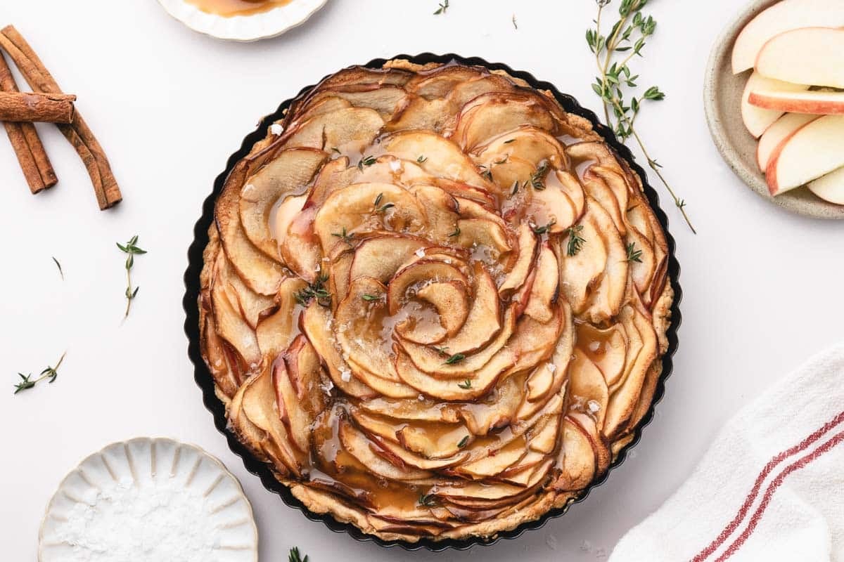 A caramel apple tart with thinly sliced apples arranged in a spiral, drizzled with caramel sauce, garnished with herbs, and surrounded by cinnamon sticks, apple slices, and a towel.