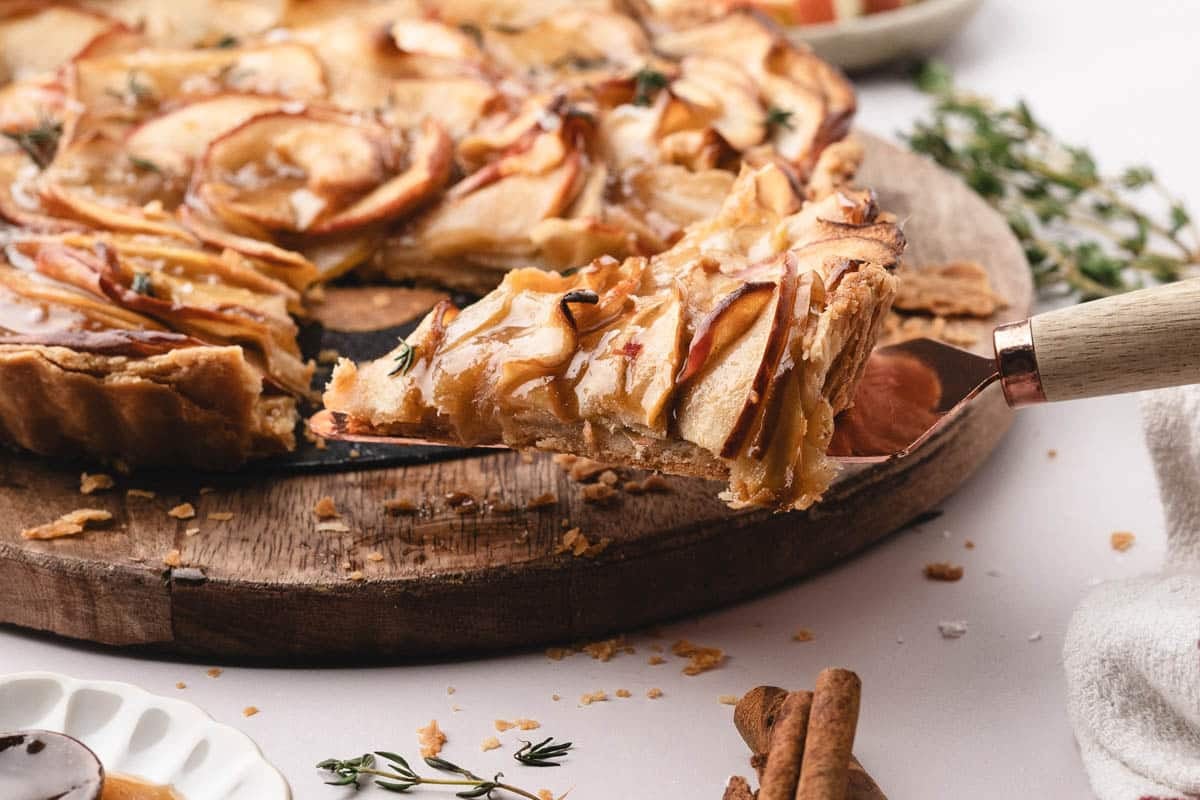 A slice of caramel apple tart being lifted from a wooden serving board, with caramel drizzle and herbs on top.