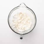 A glass mixing bowl containing flour and small lumps of butter on a white background.