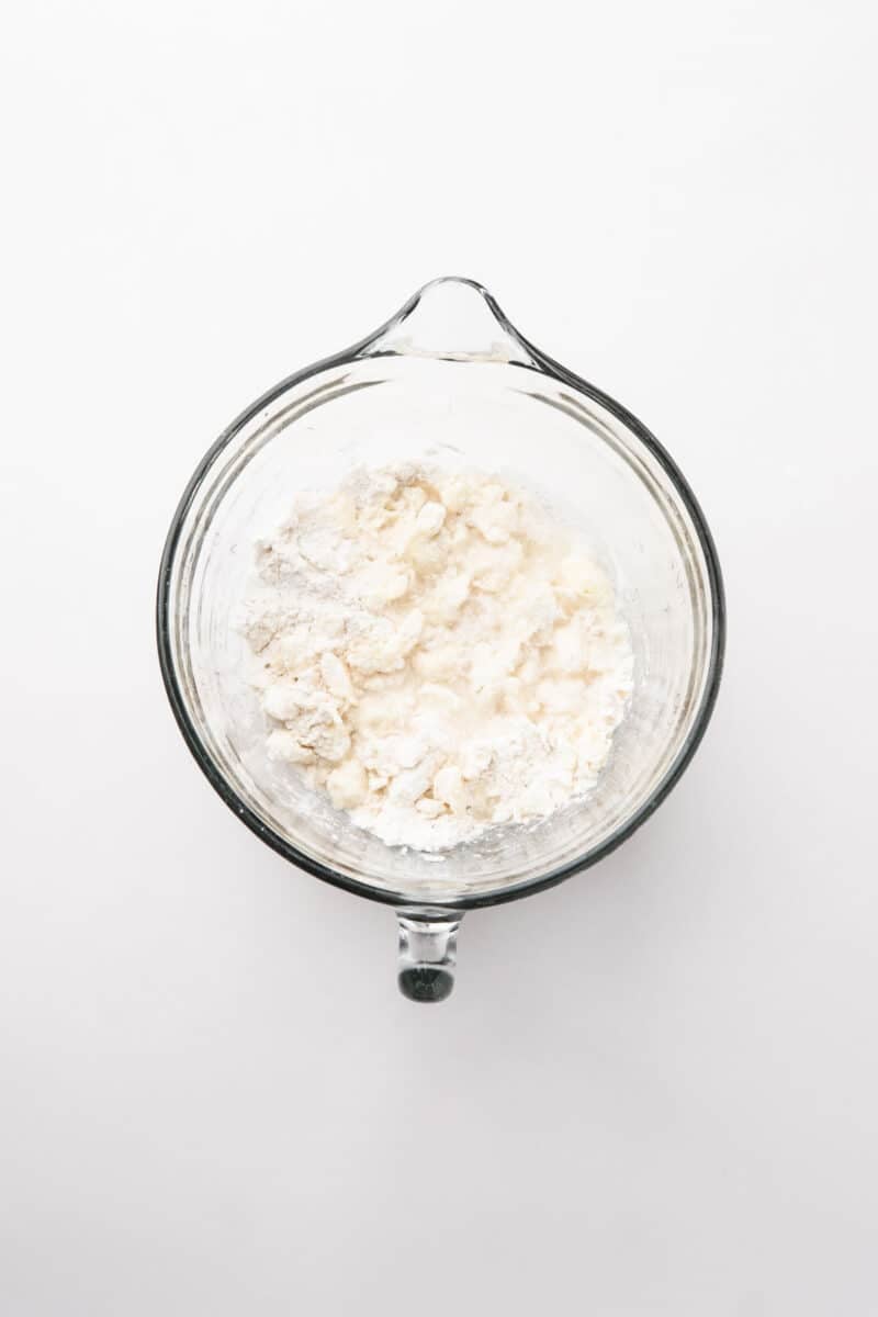 A glass mixing bowl containing flour and small lumps of butter on a white background.
