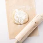 A ball of dough dusted with flour sits on parchment paper next to a wooden rolling pin on a white surface.
