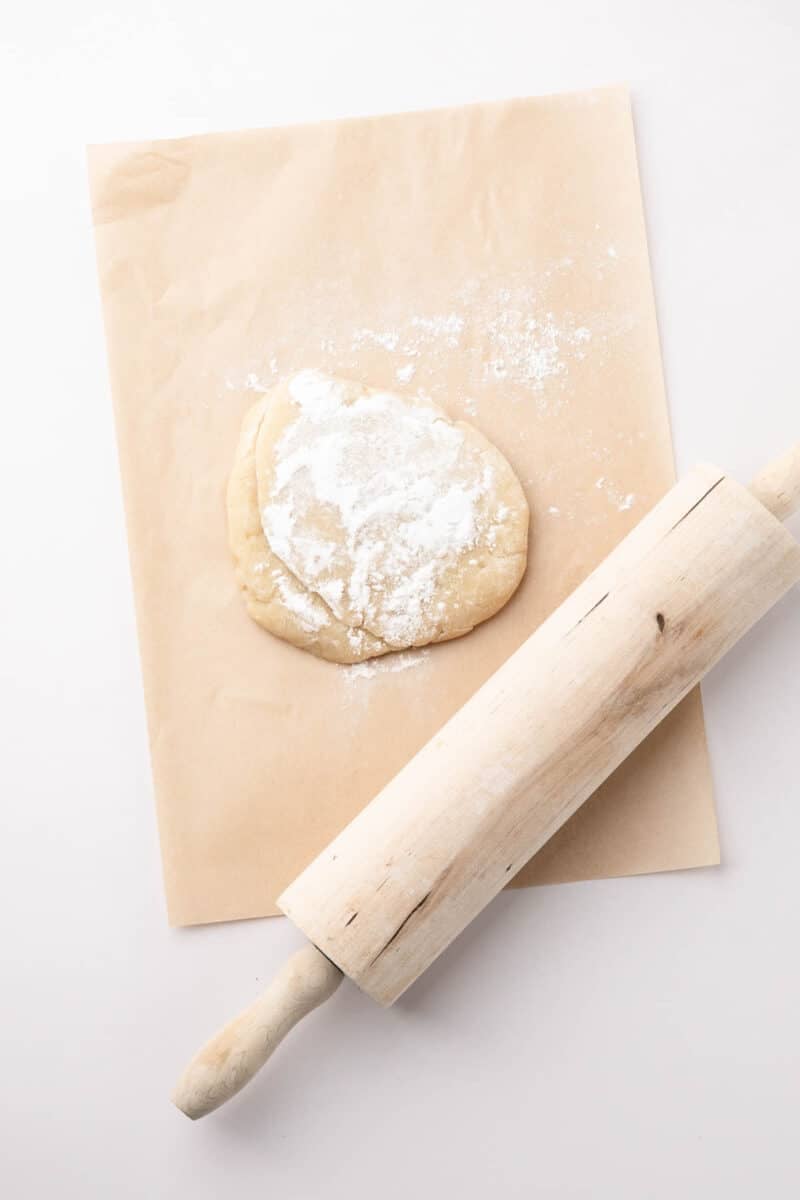 A ball of dough dusted with flour sits on parchment paper next to a wooden rolling pin on a white surface.