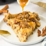 Two slices of caramel pecan roll topped with pecans are drizzled with caramel sauce on a white plate, next to a gold fork and cinnamon sticks.