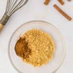 A glass bowl with brown sugar and ground cinnamon, next to a whisk and three whole cinnamon sticks on a white surface.