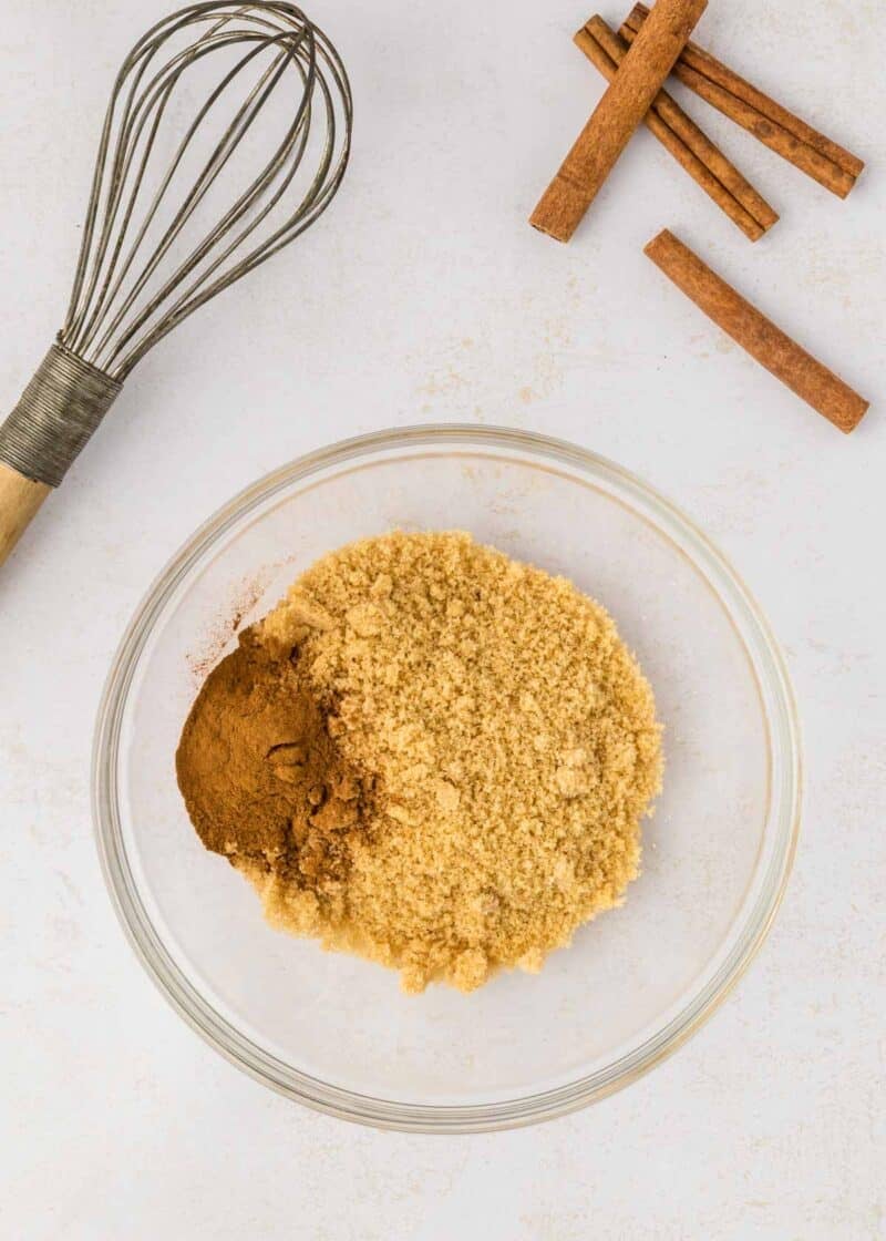 A glass bowl with brown sugar and ground cinnamon, next to a whisk and three whole cinnamon sticks on a white surface.