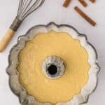 A bundt pan filled with cake batter sits on a white surface next to a metal whisk and three cinnamon sticks.