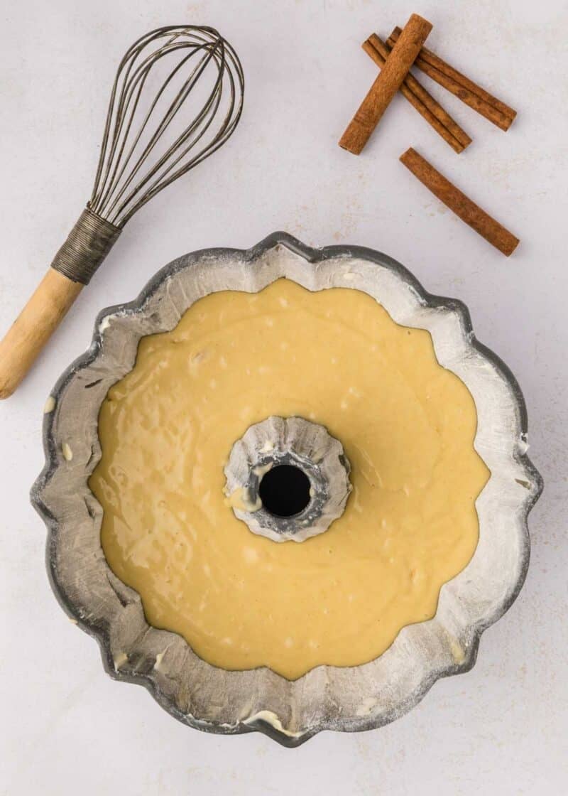 A bundt pan filled with cake batter sits on a white surface next to a metal whisk and three cinnamon sticks.