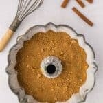 A bundt pan filled with brown sugar mixture sits on a white surface next to a whisk and three cinnamon sticks.