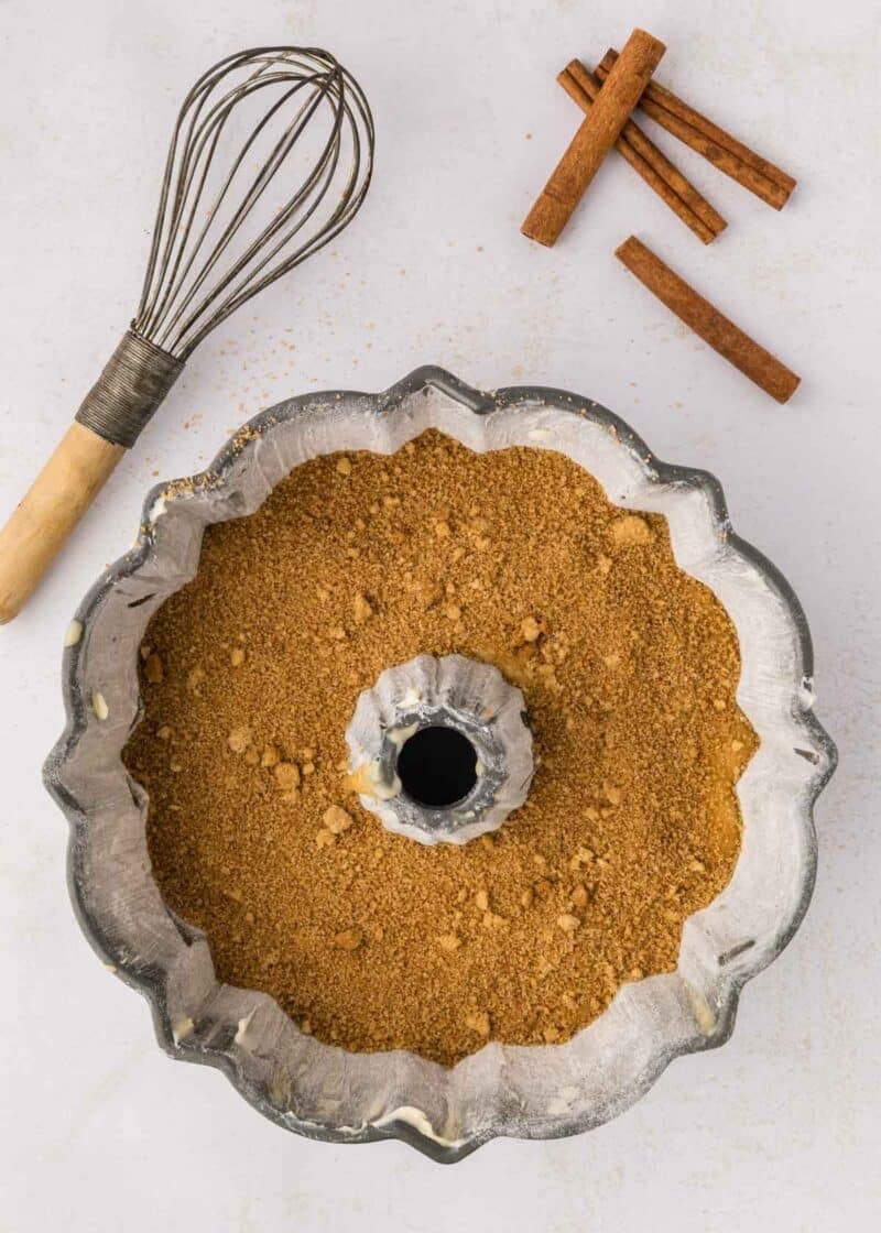 A bundt pan filled with brown sugar mixture sits on a white surface next to a whisk and three cinnamon sticks.