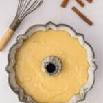 Bundt cake pan filled with batter sits on a light surface next to a whisk and three cinnamon sticks.