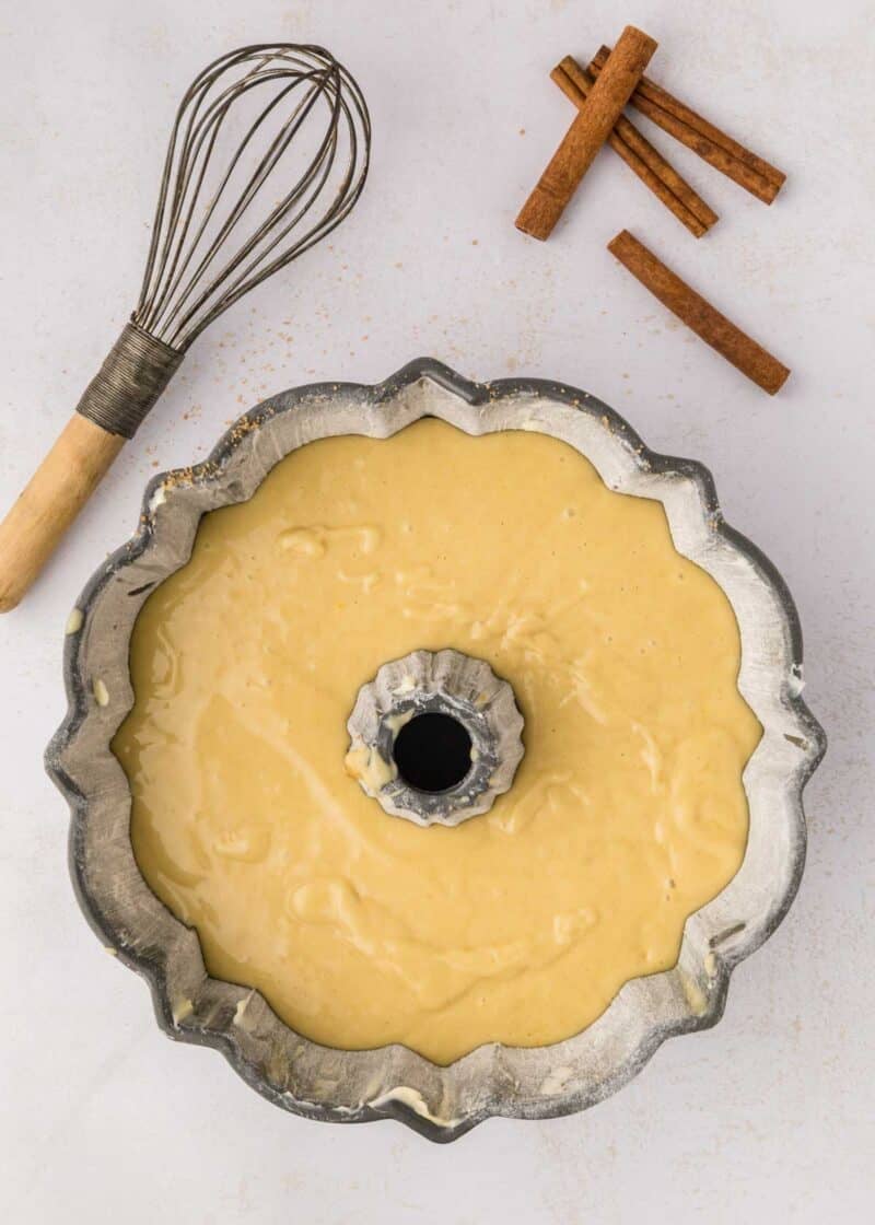 Bundt cake pan filled with batter sits on a light surface next to a whisk and three cinnamon sticks.