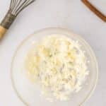 A glass bowl with creamed butter, a metal whisk, and cinnamon sticks on a light surface.