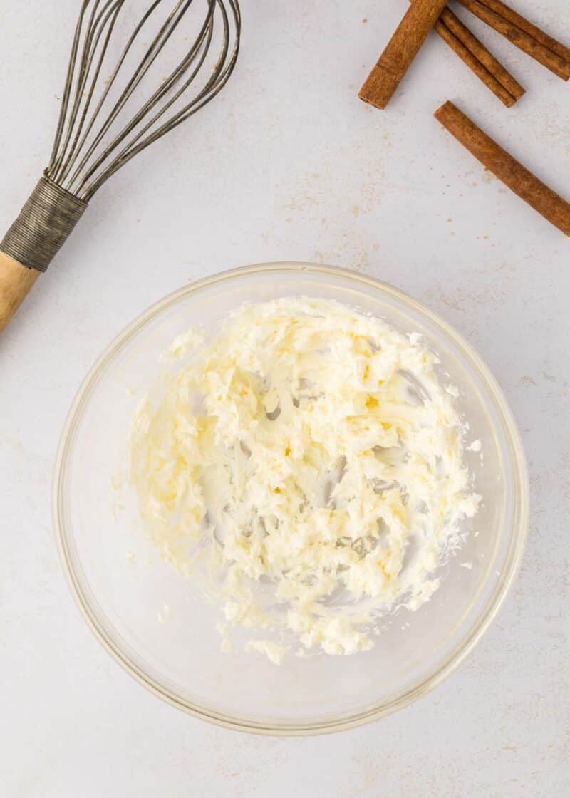 A glass bowl with creamed butter, a metal whisk, and cinnamon sticks on a light surface.