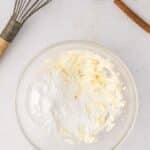 A glass bowl with butter and powdered sugar sits on a white surface near a whisk and several sticks of cinnamon.