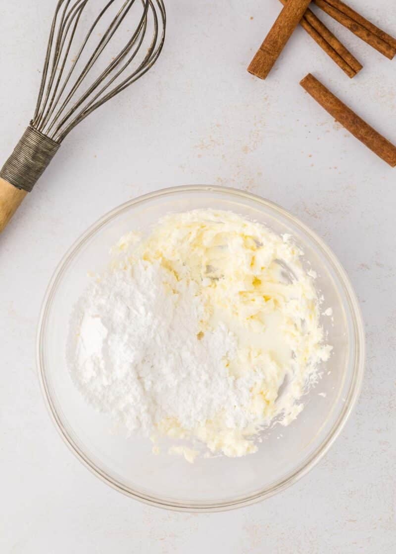 A glass bowl with butter and powdered sugar sits on a white surface near a whisk and several sticks of cinnamon.