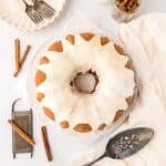 A cinnamon bundt cake with white icing is on a plate, surrounded by forks, plates, a cake server, cinnamon sticks, a grater, and a jar of milk on a light-colored table.