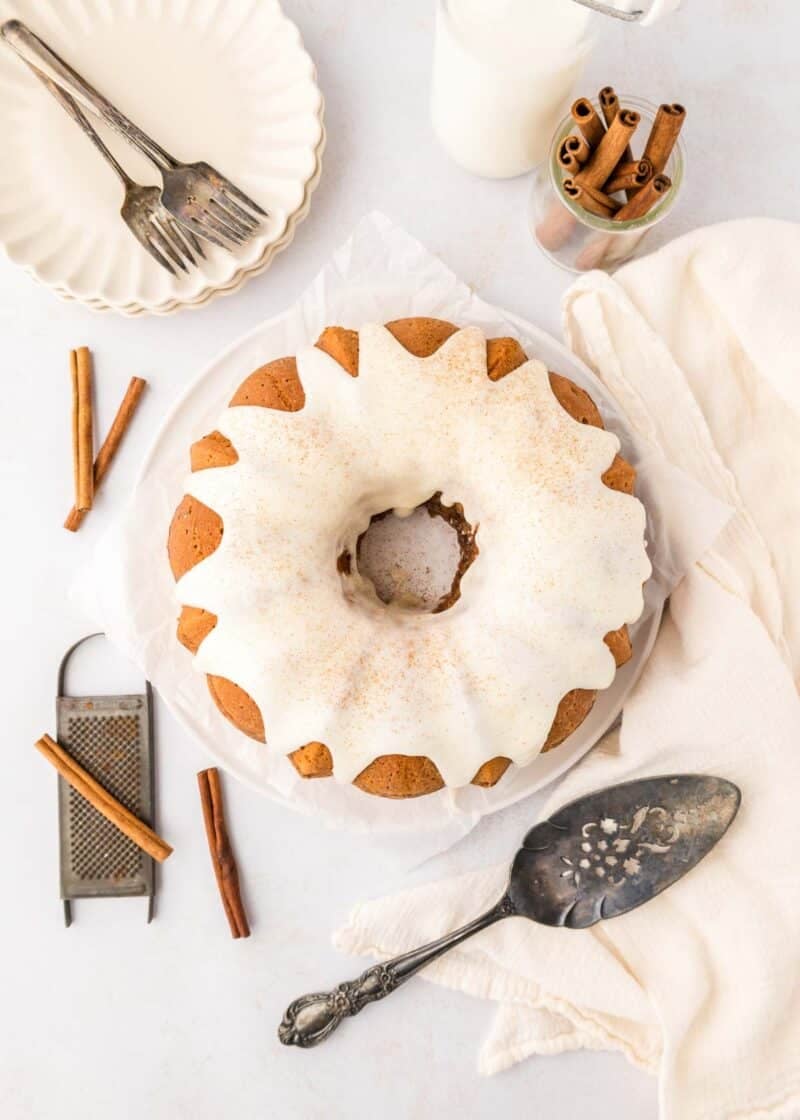 A cinnamon bundt cake with white icing is on a plate, surrounded by forks, plates, a cake server, cinnamon sticks, a grater, and a jar of milk on a light-colored table.