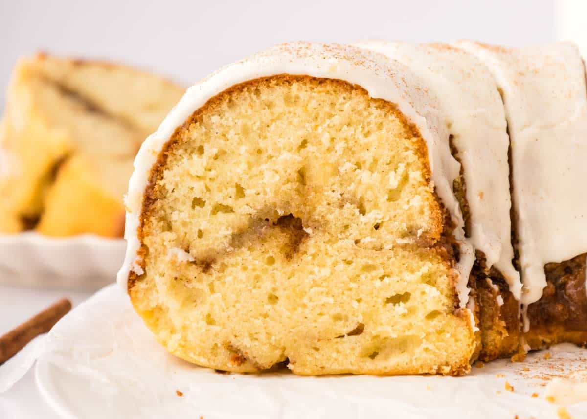 Close-up of a sliced bundt cake with white icing on top, showing a moist, light yellow interior and swirls of cinnamon.