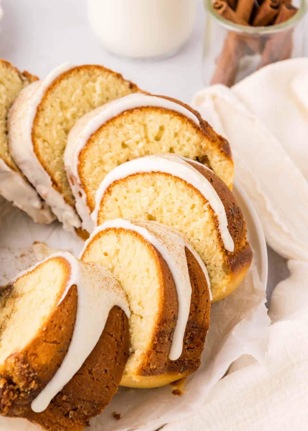 Sliced cinnamon bundt cake with white icing drizzled on top, arranged on parchment paper, with a jar of cinnamon sticks and a glass of milk in the background.