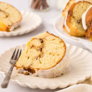 A slice of cinnamon swirl bundt cake with icing on a white plate, a fork beside it, and more cake in the background.