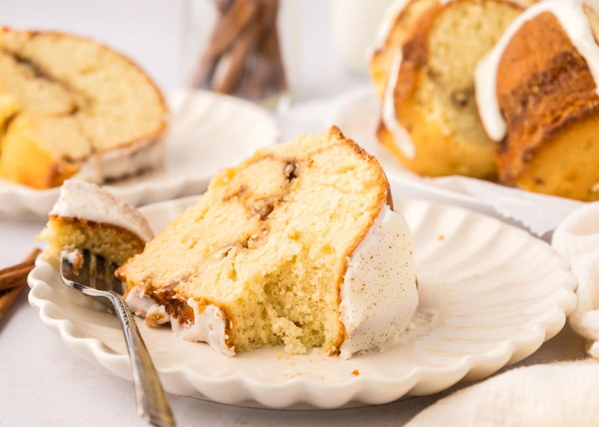 A slice of frosted bundt cake with a fork on a white plate, with more cake and cinnamon sticks visible in the background.