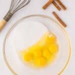 A glass bowl with six cracked eggs and sugar, next to a whisk and several sticks of cinnamon on a light surface.