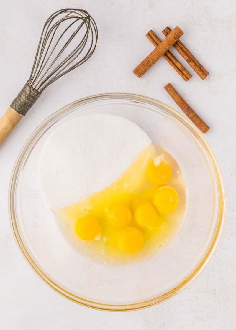 A glass bowl with six cracked eggs and sugar, next to a whisk and several sticks of cinnamon on a light surface.