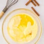 A glass bowl with whisked eggs, milk, and melted butter on a white surface, next to a whisk and several cinnamon sticks.