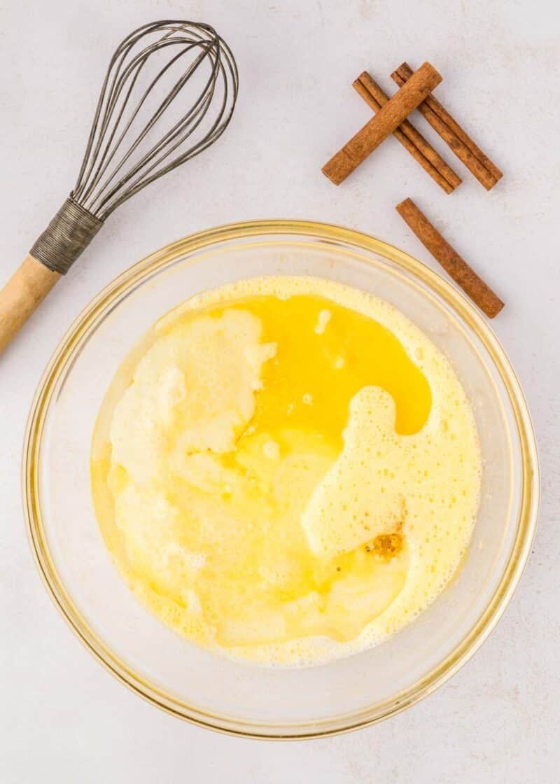 A glass bowl with whisked eggs, milk, and melted butter on a white surface, next to a whisk and several cinnamon sticks.