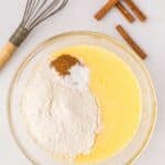 A glass bowl with beaten eggs, flour, baking powder, and spices sits on a counter next to a whisk and cinnamon sticks.