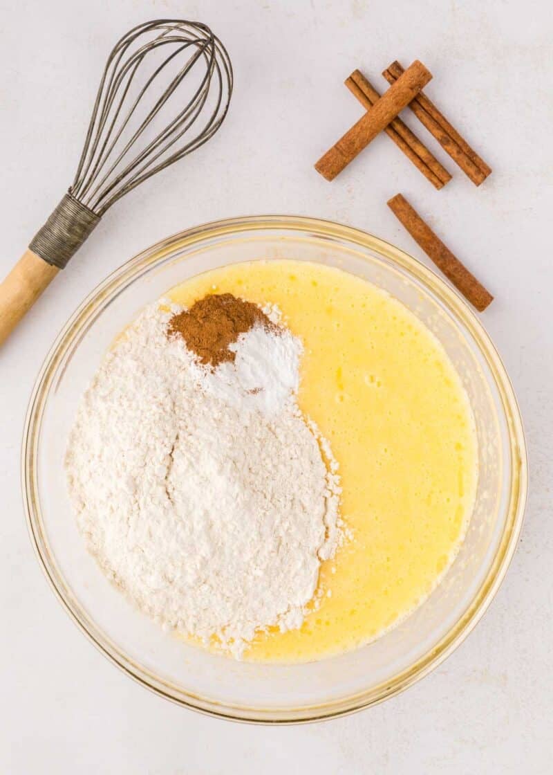 A glass bowl with beaten eggs, flour, baking powder, and spices sits on a counter next to a whisk and cinnamon sticks.