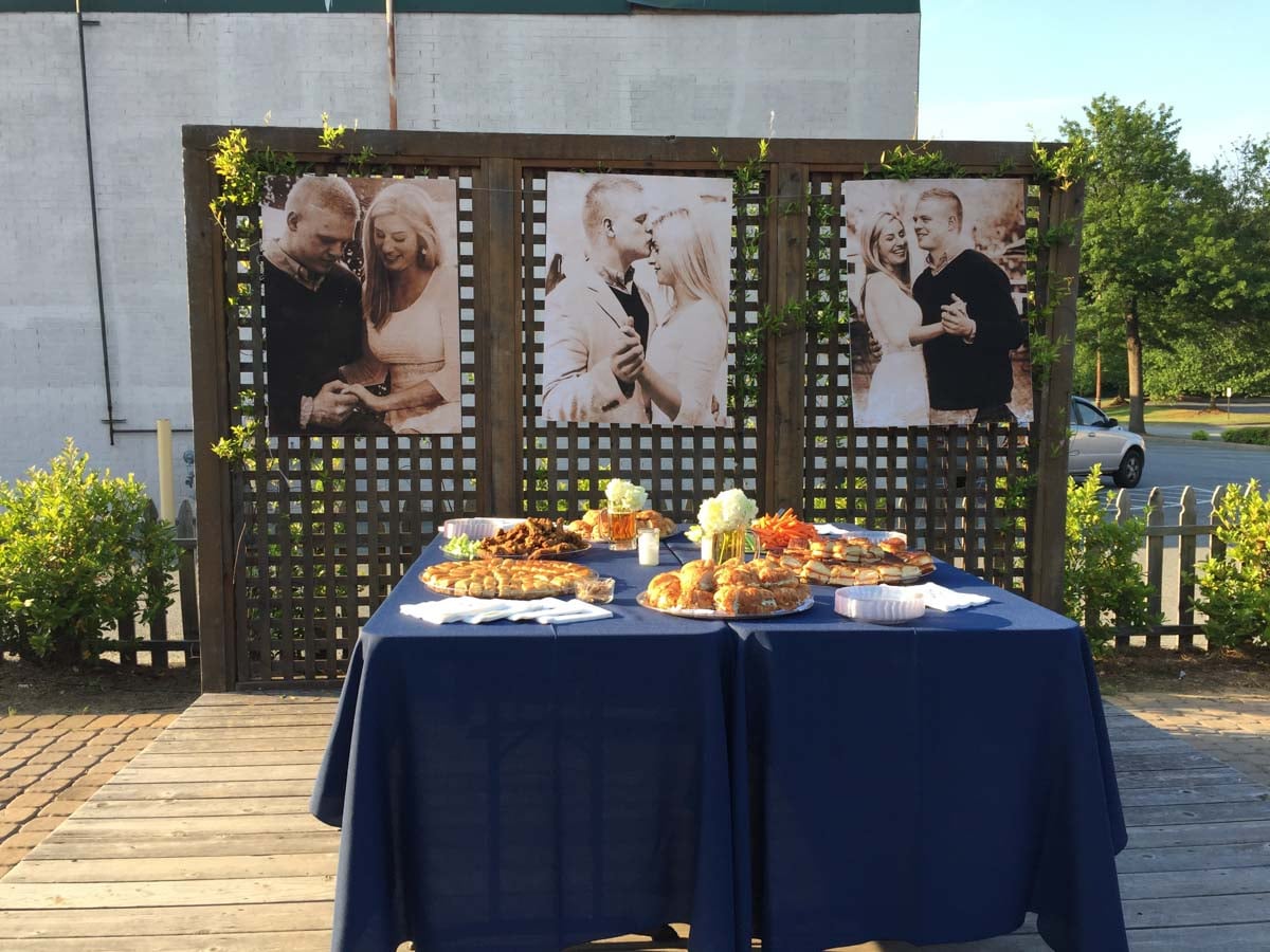 A table with pastries and flowers sits in front of a wooden lattice fence displaying three large black-and-white couple photos at an outdoor event.
