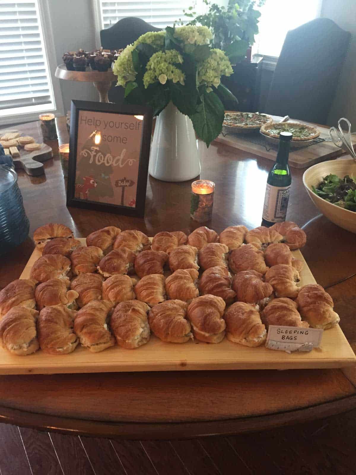 A wooden tray of croissant sandwiches labeled “Sleeping Bags” on a dining table, surrounded by salad, drinks, cupcakes, and a decorative flower arrangement.