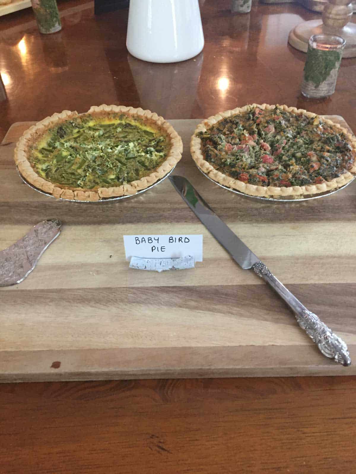 Two savory pies sit on a wooden board with a silver knife between them; a small sign in front reads “BABY BIRD PIE.”.