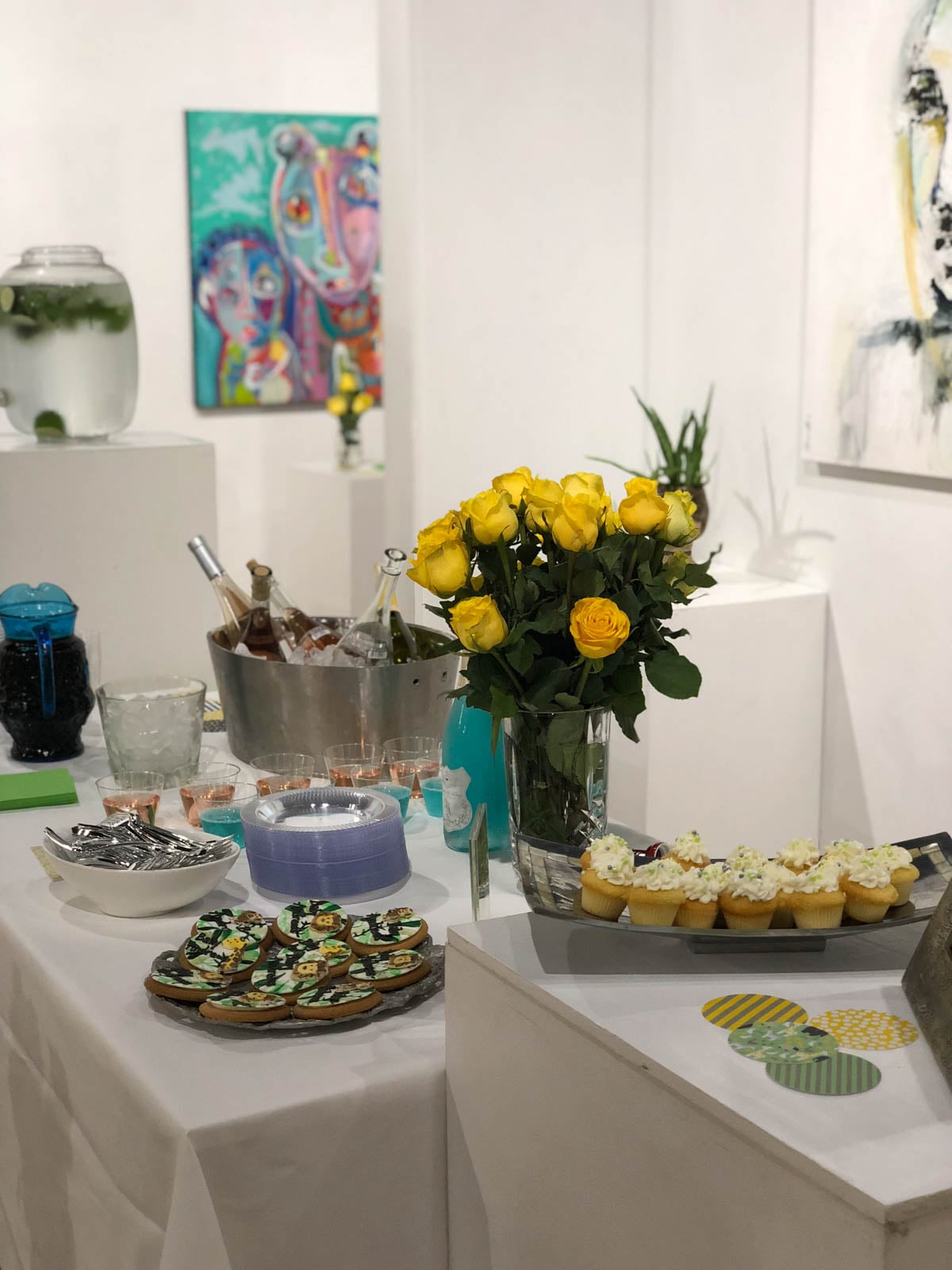 Table set with yellow roses, decorated cupcakes, cookies, drinks, and utensils in an art gallery with colorful paintings on the walls.