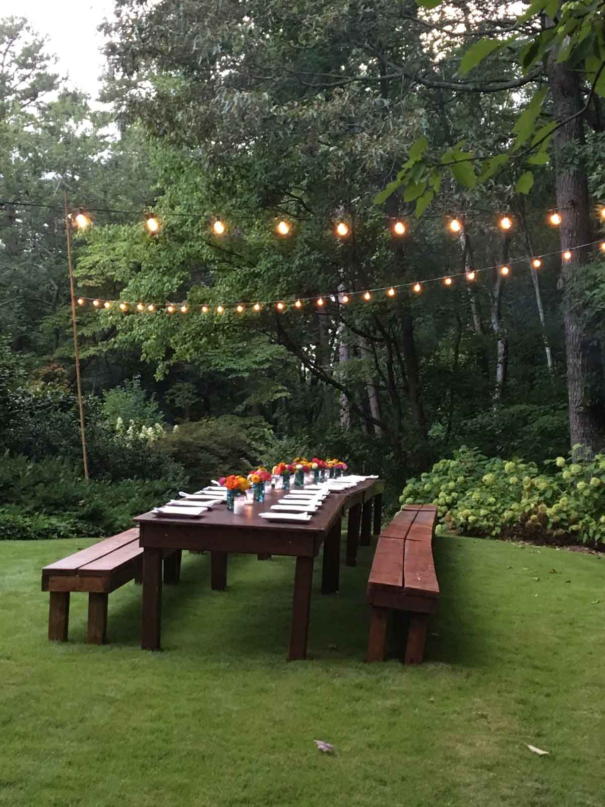 Long wooden picnic table set with plates and flowers, surrounded by benches and string lights, in a grassy yard with trees in the background.