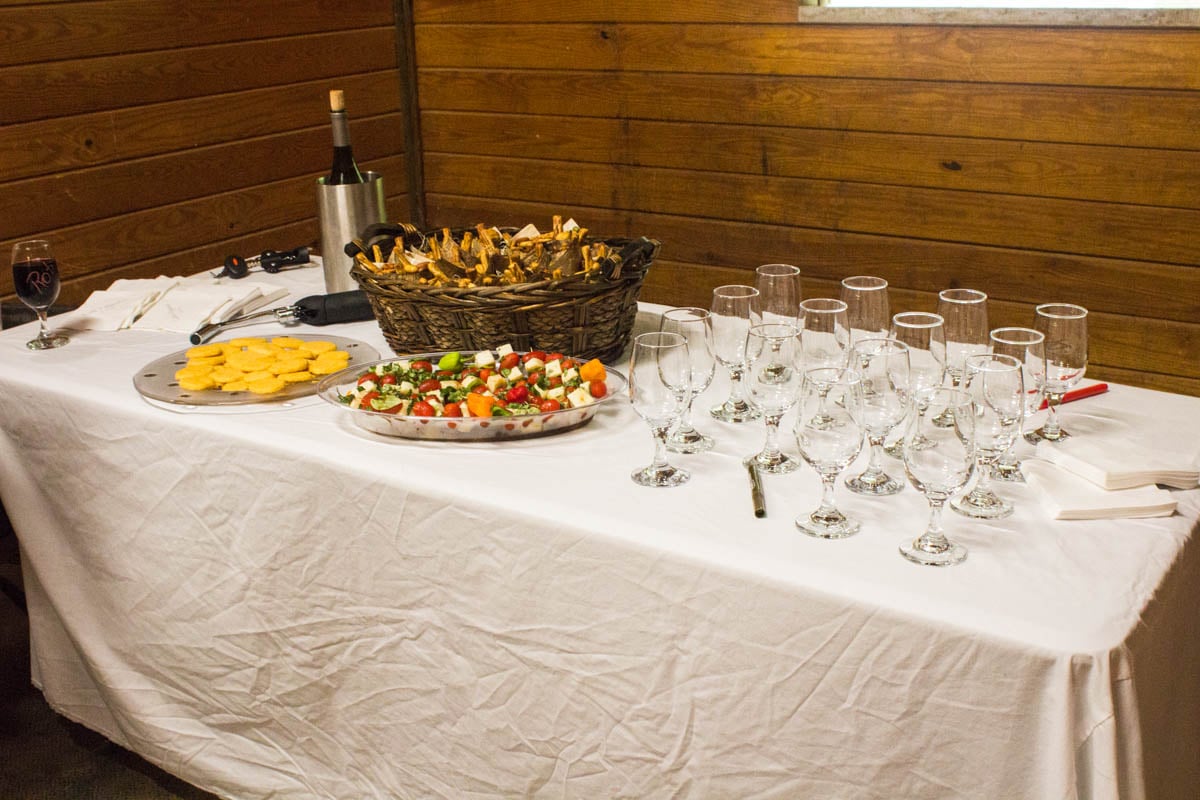 A table with wine glasses, a wine bottle, a basket of breadsticks, a plate of crackers, and a platter of skewered appetizers on a white tablecloth.