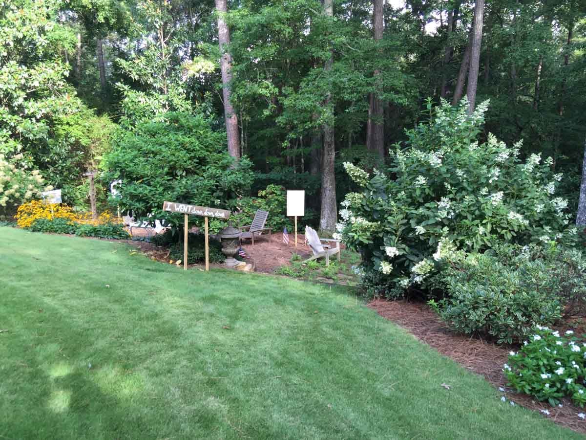 A grassy lawn with a small garden seating area featuring wooden chairs, lush green bushes, flowering plants, and a sign reading “Visitor Lounge.” Tall trees fill the background.
