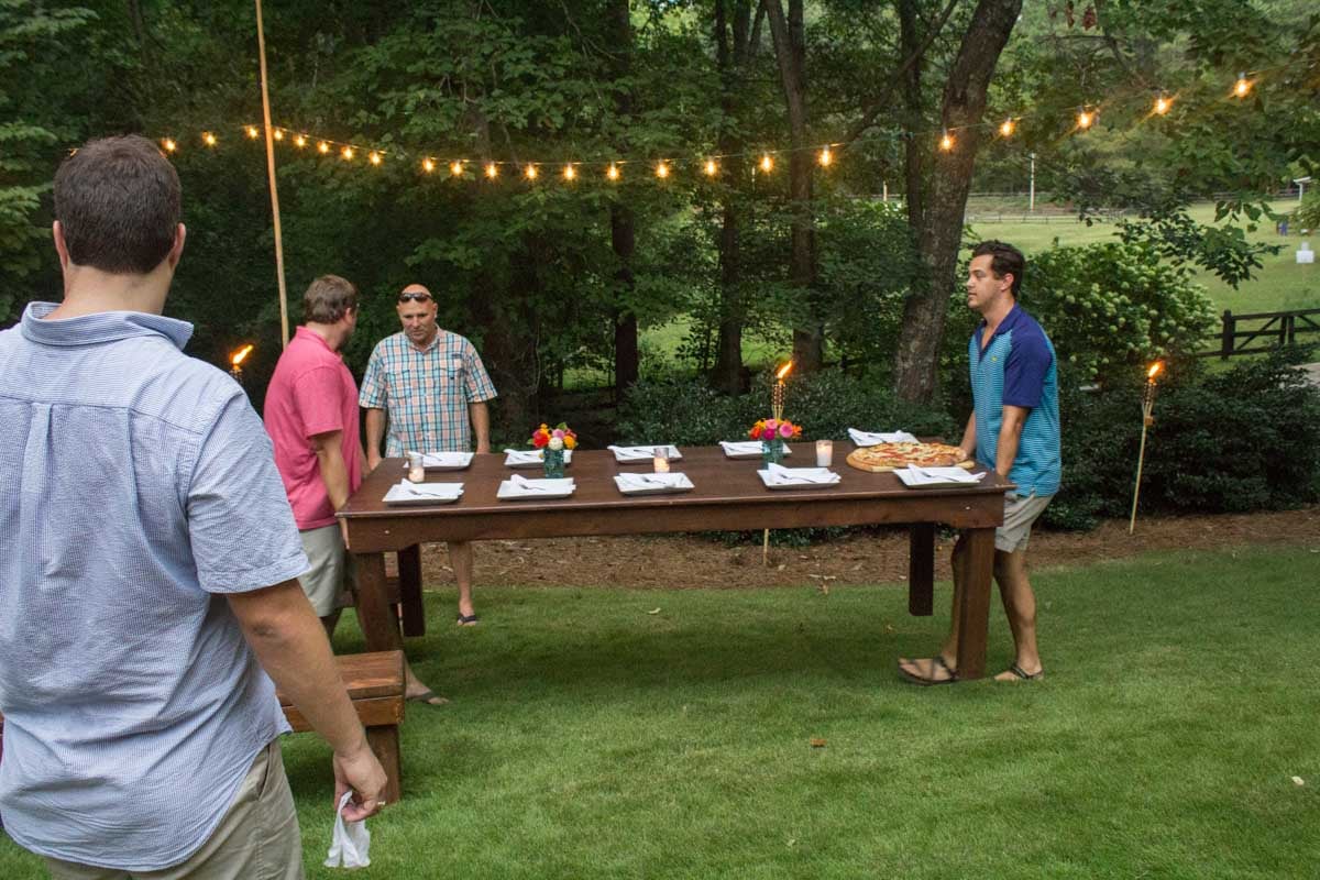 Four men stand around an outdoor table set with plates, napkins, and flowers under string lights on a grassy lawn surrounded by trees.
