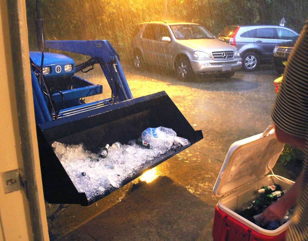 A tractor bucket filled with ice and drinks sits in a driveway during heavy rain; a person adds drinks to a cooler in the foreground.