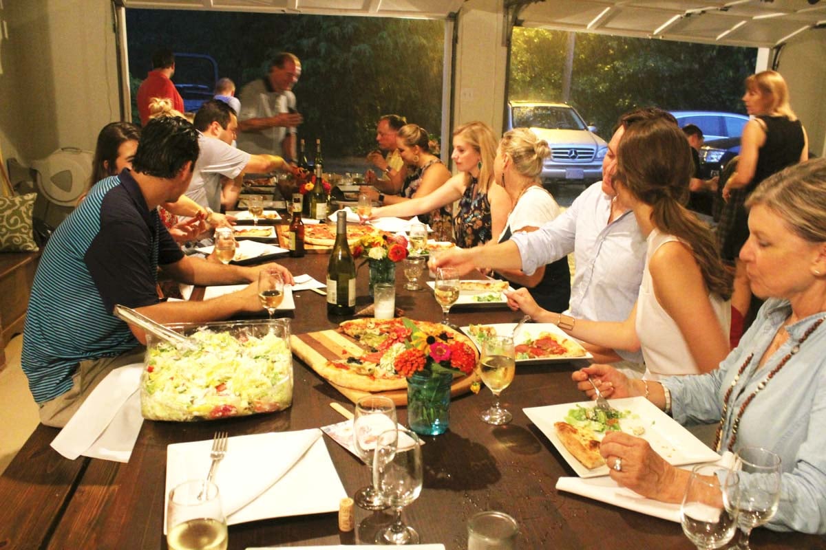A group of people are seated around a large dining table, eating and talking, with plates of food, wine glasses, and salad visible.