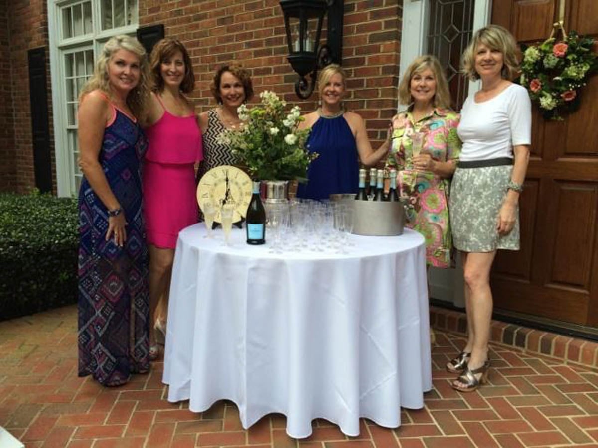Six women stand outside a house next to a table with drinks, glasses, and flowers, smiling at the camera.