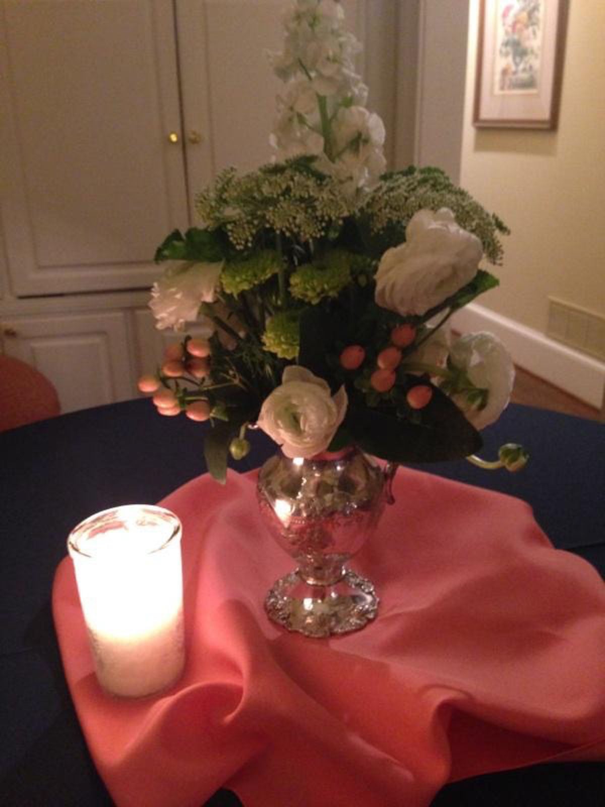 A silver vase with white flowers and pink berries sits on a coral cloth next to a lit candle on a dark table.